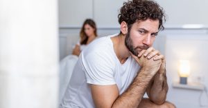 Sad thoughtful man after arguing with girlfriend.Relationship difficulties, conflict and family concept,unhappy couple having problems at bedroom. Sad guy sitting on bed,girlfriend in the background; blog: possible causes of erectile dysfunction