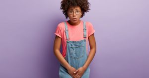 Dissatisfied girl keeps hands on crotch, presses lower abdomen, needs toilet badly, has syndrome of cystitis, wears spectacles, pink t shirt and denim sarafan, isolated on purple wall. Health problem; blog: Are There Different Types of Urinary Incontinence?